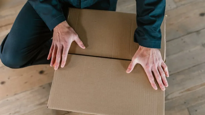 Hands closing a cardboard moving box on a wooden floor